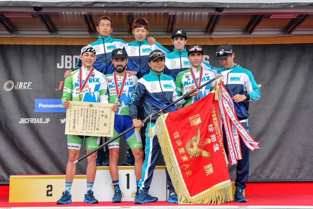 表彰台で優勝旗を掲げるチーム集合写真